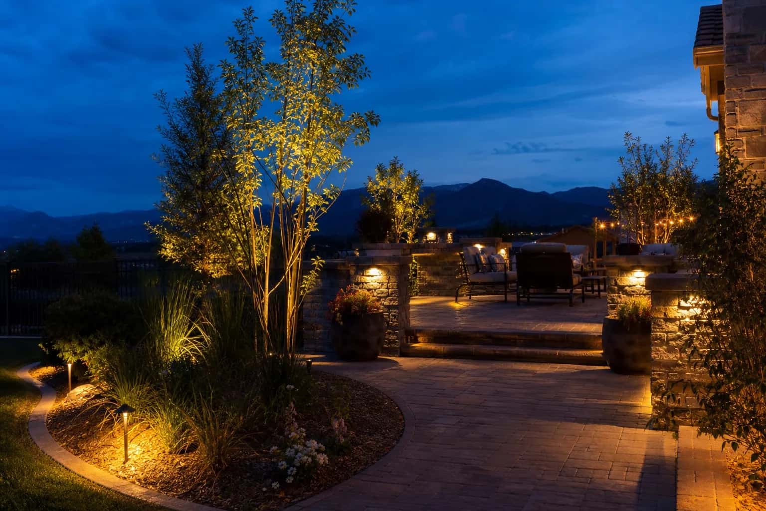 Paver patio at twilight with warm path lights, uplighting on stone columns and trees, low-voltage outdoor lighting scene
