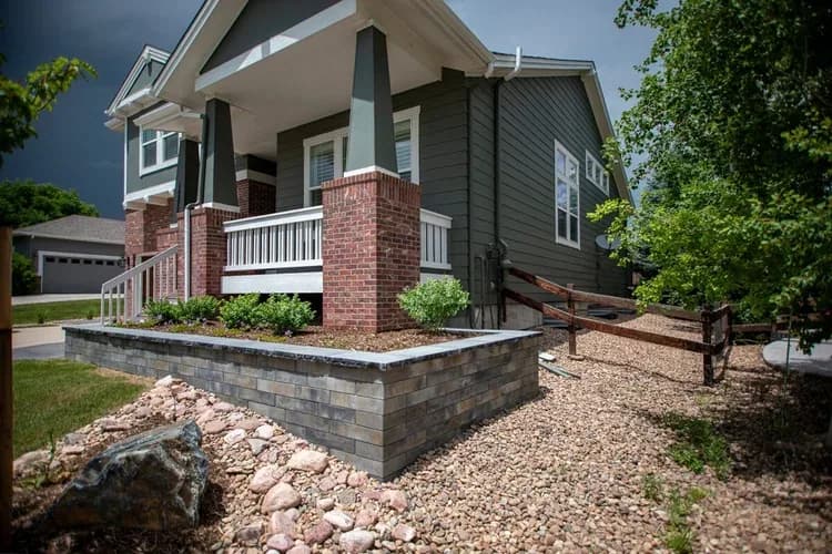 Craftsman porch with white railings, brick column bases, capped stone planter, and gravel transition to the lawn.