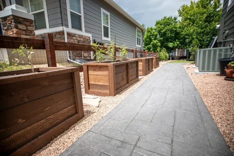 Side yard: stamped concrete walk, stained cedar raised beds on pea gravel, and stone veneer at the base of the grey-sided home.