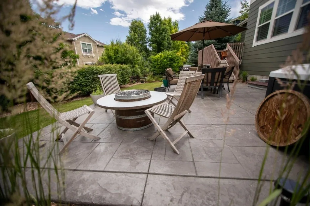 Sorensen Westminster backyard: stamped concrete patio with barrel-style fire table, wooden sling chairs, dining zone under umbrella, and olive-toned siding