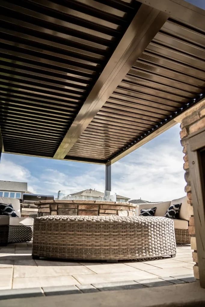 Bronze powder-coated louvered patio cover over tan pavers, circular stone fire pit with glass guard, and wicker seating, Colorado outdoor living