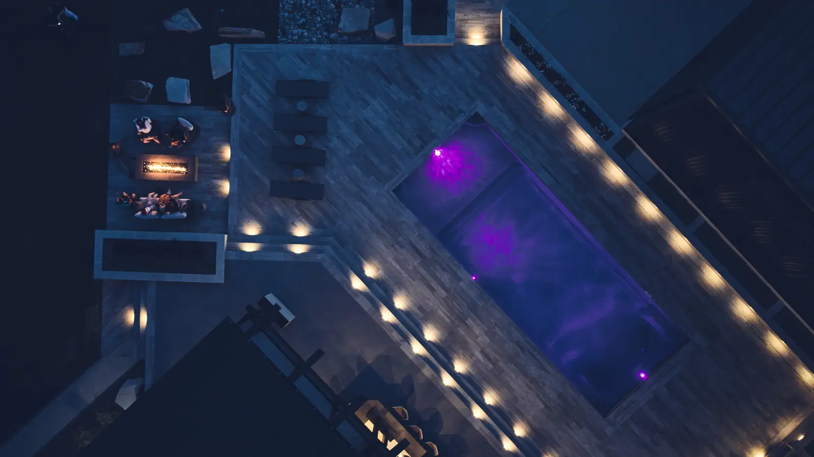 Night aerial of an LED-lit pool glowing purple beside a burning fire pit lounge