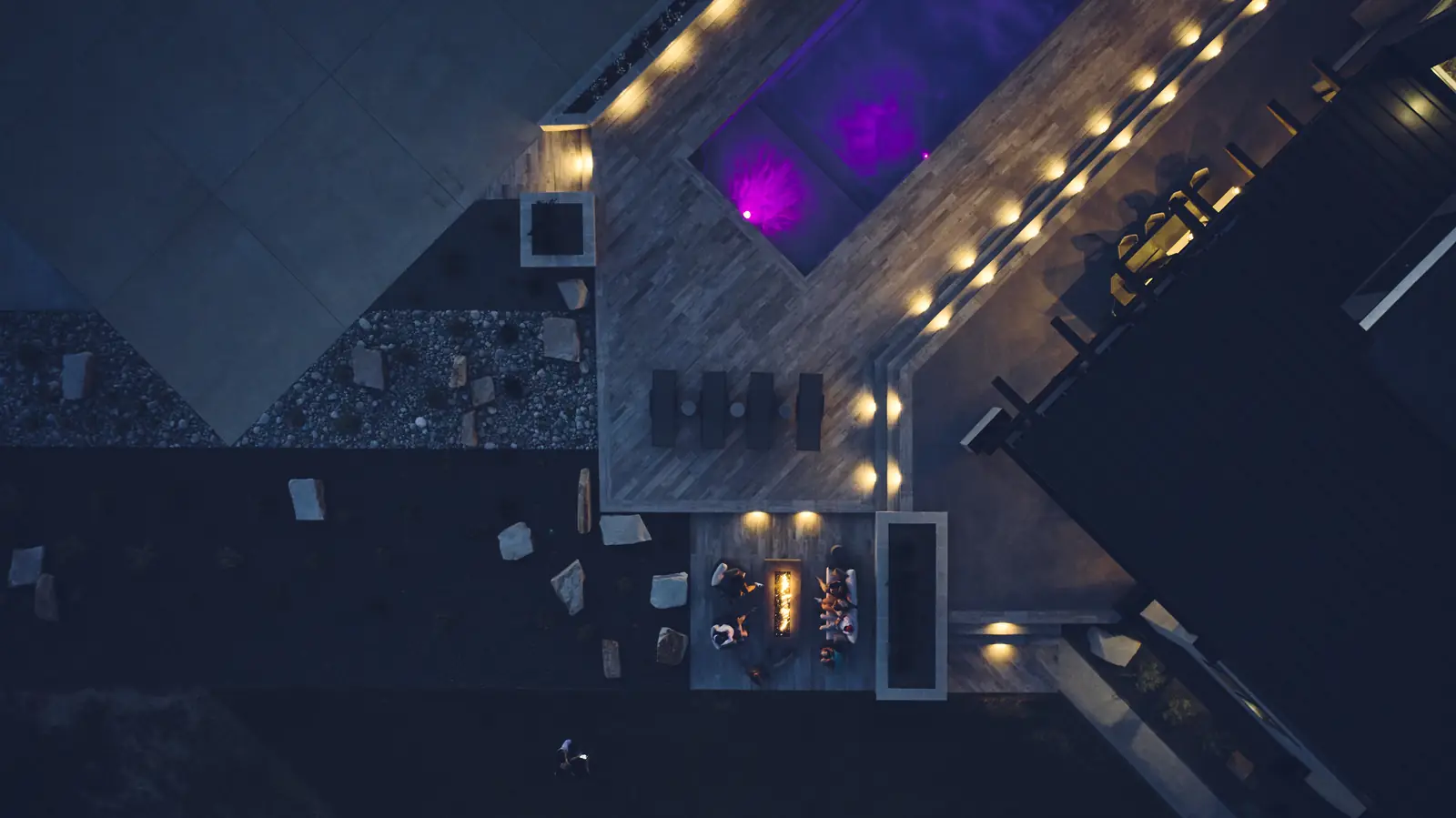 Night aerial of a purple-lit pool and spa with deck step lighting and fire pit